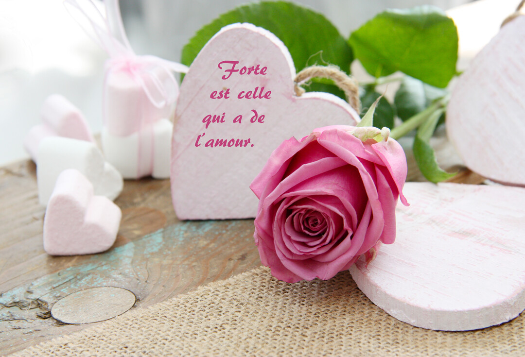 Une rose rose est posée près de cœurs en bois sur une table rustique, l'un d'eux portant une citation sur l'amour et la force.