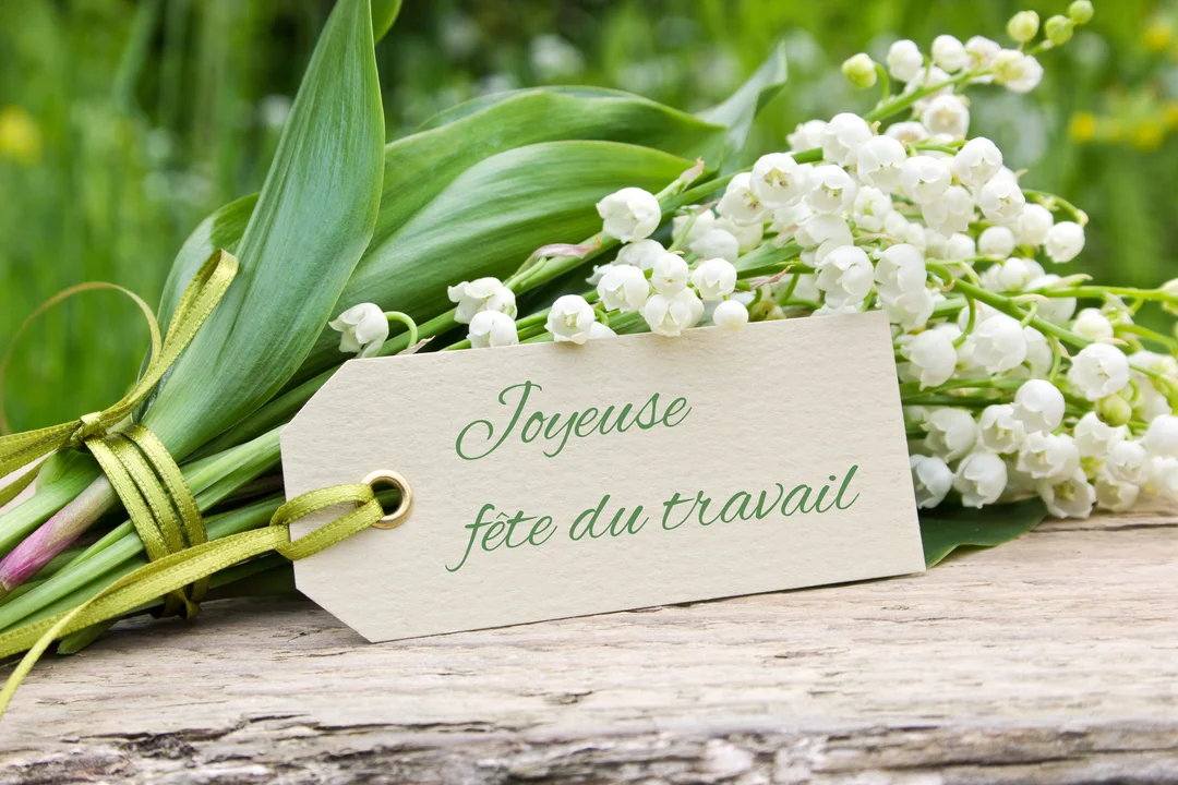Une photographie en gros plan d'un bouquet de muguet aux clochettes blanches, posé sur du bois rustique avec une étiquette souhaitant une joyeuse fête du travail.