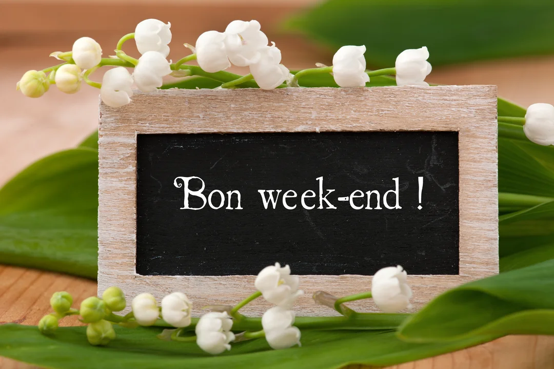 Une ardoise rustique affichant 'Bon week-end !' est posée sur des brins de muguet frais aux clochettes blanches et feuilles vertes.