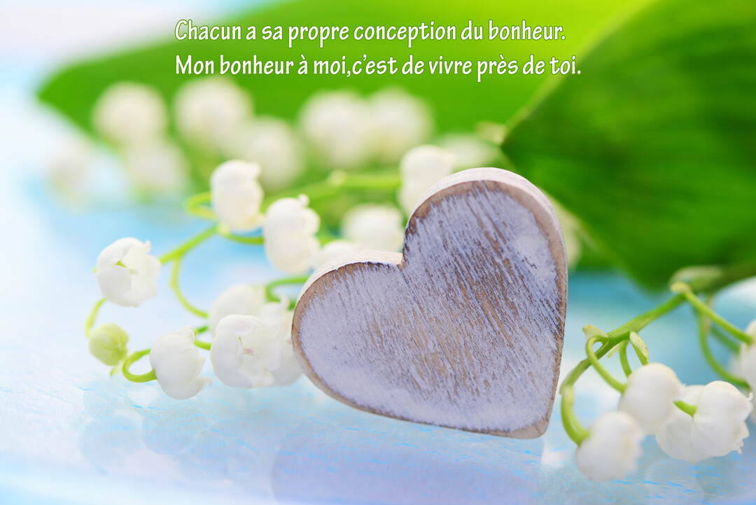 Un cœur en bois blanc est posé devant des brins de muguet en fleurs sur un fond bleu et vert doux et flouté.