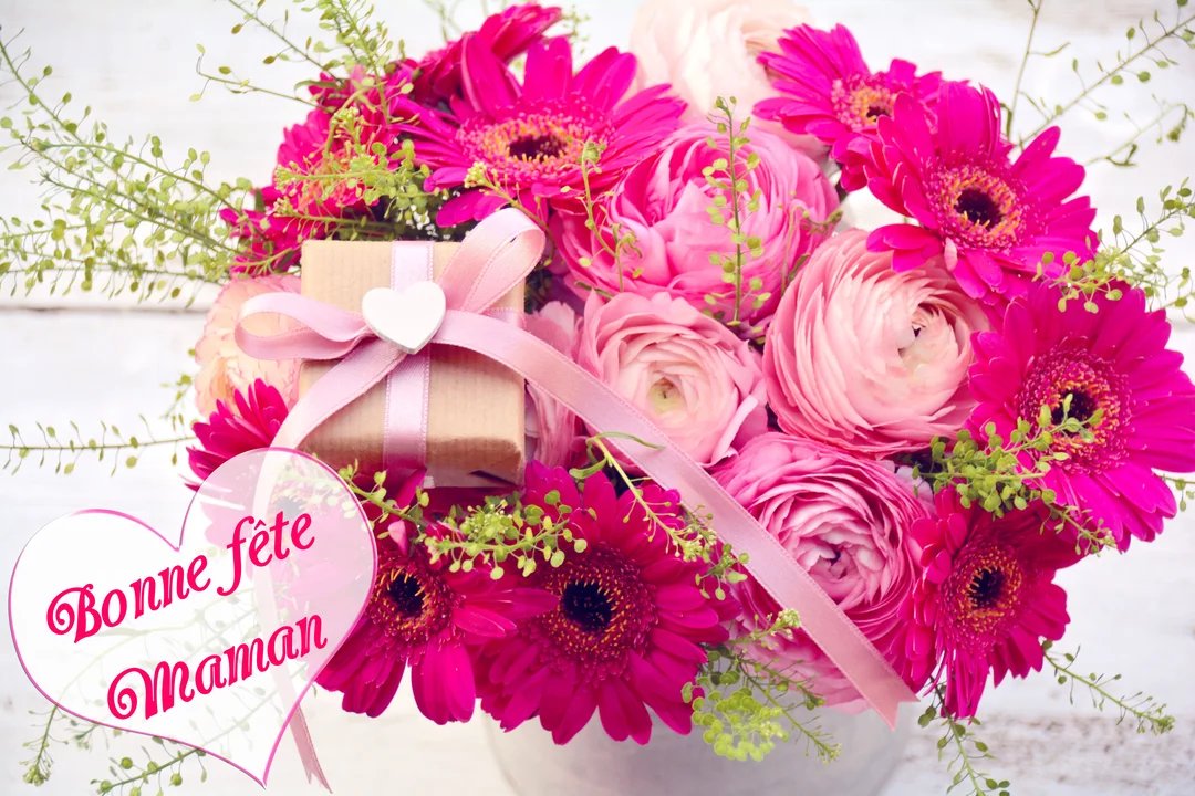 Un somptueux bouquet de fleurs roses et fuchsia, accompagné d'un petit cadeau, sur un fond en bois blanc avec l'inscription "Bonne fête maman".