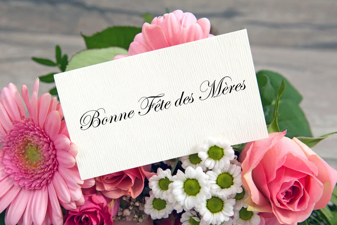 Une carte de vœux blanche avec l'inscription 'Bonne Fête des Mères' est posée sur un bouquet de fleurs fraîches roses et blanches.