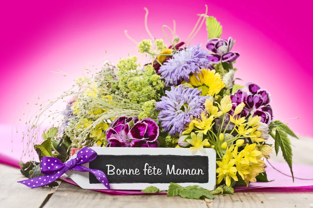 Un bouquet de fleurs sauvages violettes, jaunes et roses est posé sur une table en bois avec une étiquette "Bonne fête Maman".