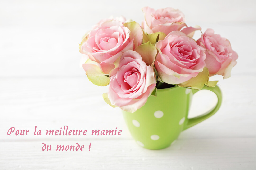 Un bouquet de roses roses délicates est présenté dans un mug vert à pois blancs, posé sur une surface en bois peinte en blanc.