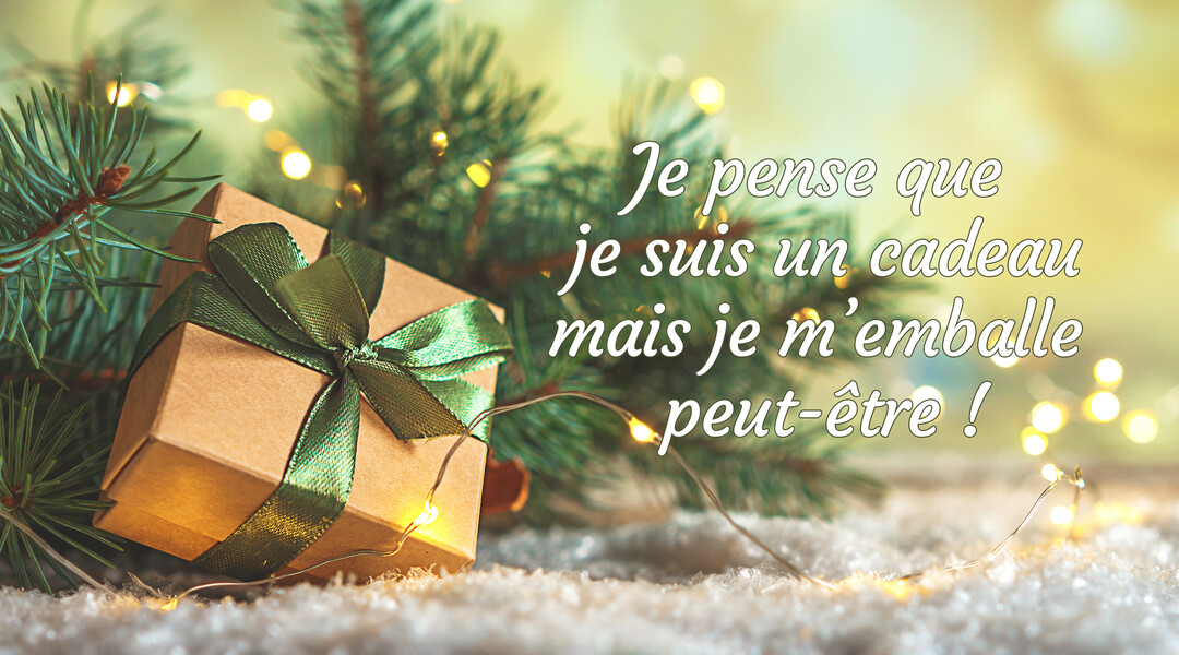 Un cadeau carré en carton avec un ruban vert est posé sur de la neige, devant des branches de sapin et une guirlande lumineuse.