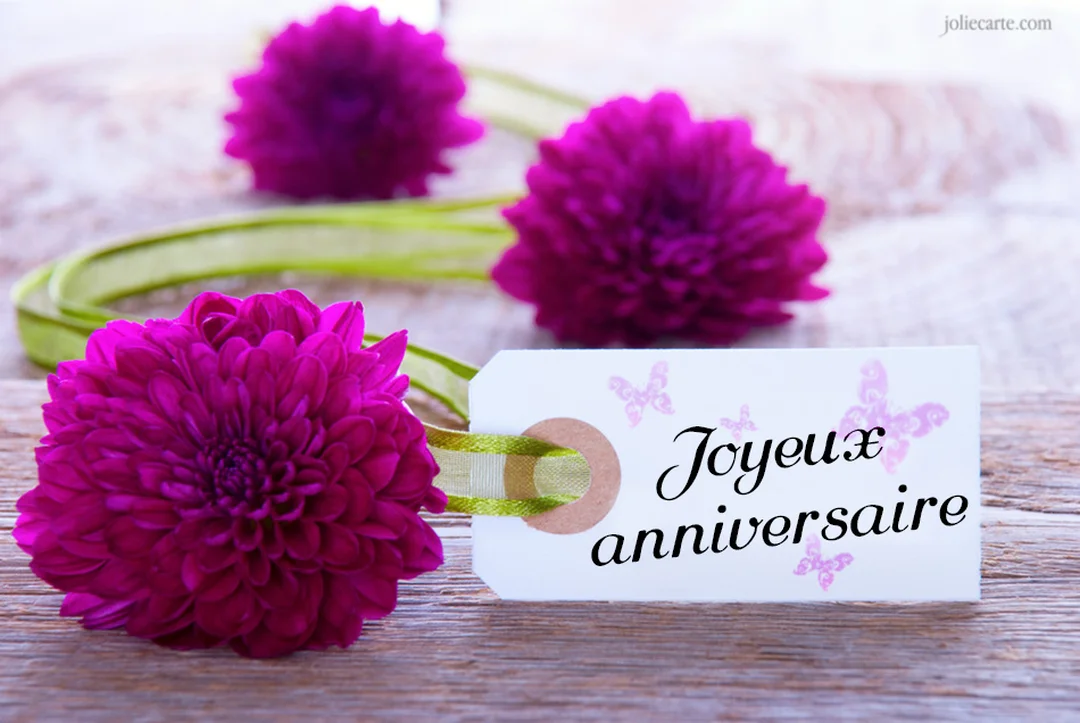 Une composition photographique montrant des fleurs magenta à côté d'une étiquette en papier sur laquelle est écrit 'Joyeux anniversaire' sur un fond en bois.