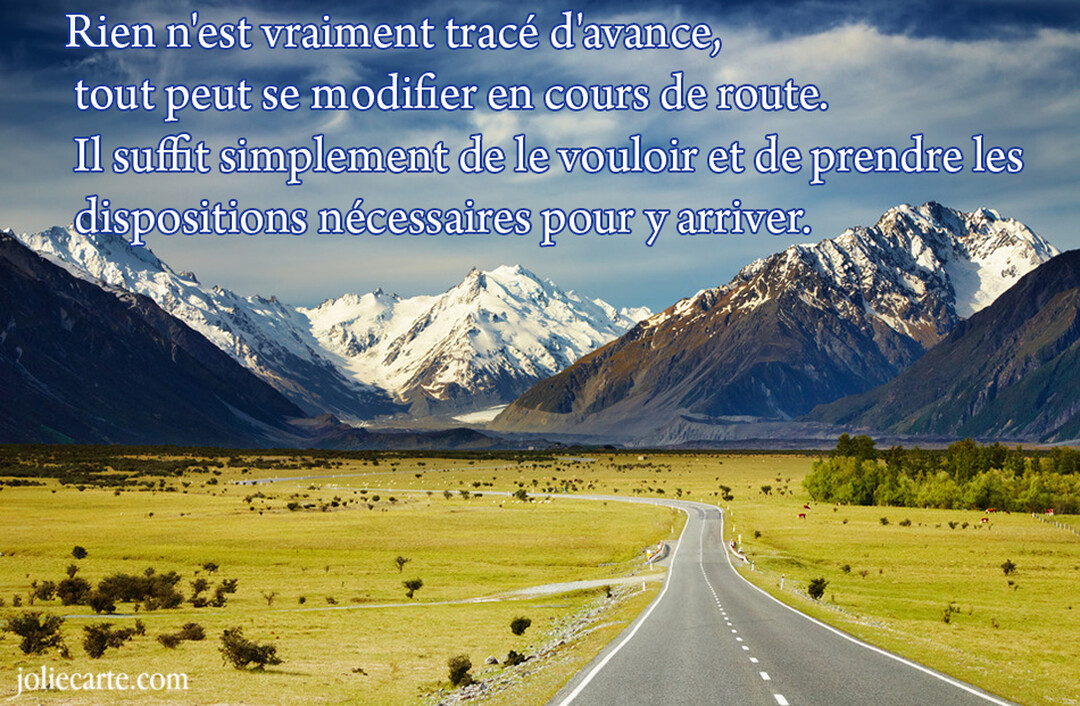 Une route asphaltée s'étire dans une vallée verdoyante vers une imposante chaîne de montagnes aux sommets enneigés, sous un ciel bleu nuageux.