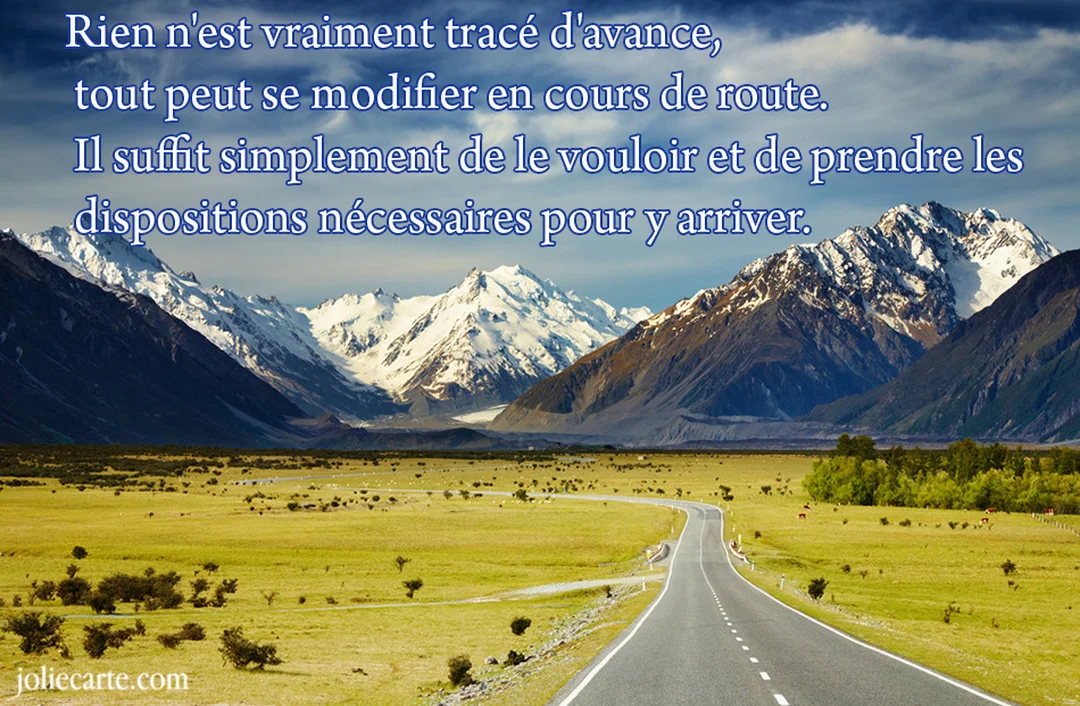 Une route asphaltée s'étire dans une vallée verdoyante vers une imposante chaîne de montagnes aux sommets enneigés, sous un ciel bleu nuageux.