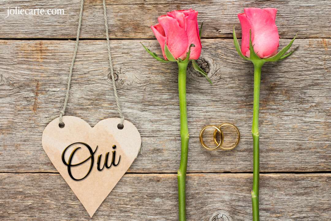 Sur un fond de bois, deux roses roses, des alliances et un cœur suspendu affichant le mot 'Oui' symbolisent une annonce de mariage.
