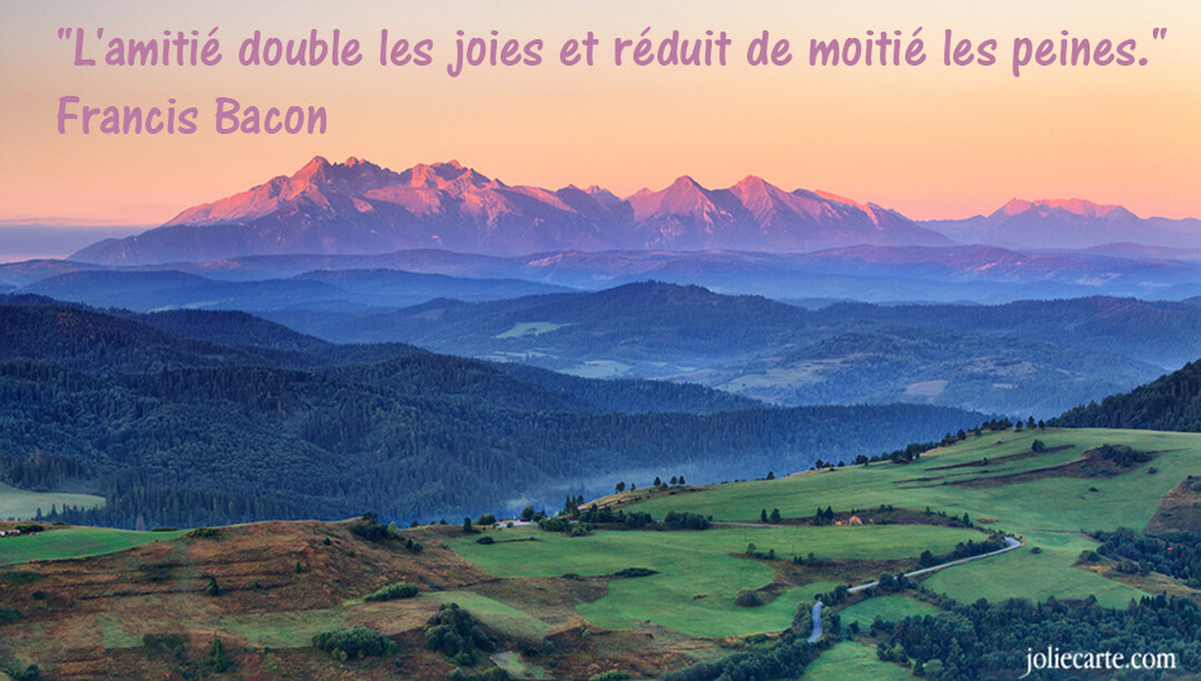 Une chaîne de montagnes aux sommets rosés par le soleil couchant surplombe des collines verdoyantes et une vallée brumeuse et bleue.