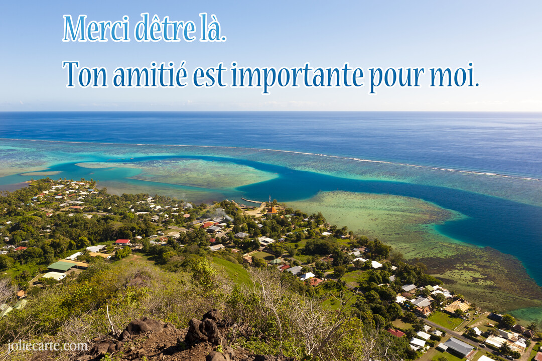 Vue en plongée d'un village côtier sur une île tropicale, bordé par un lagon turquoise et un vaste océan sous un ciel bleu.