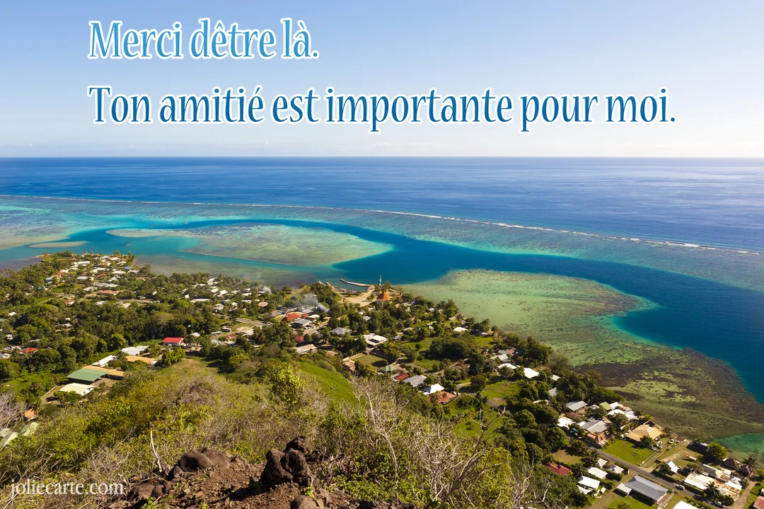 Vue en plongée d'un village côtier sur une île tropicale, bordé par un lagon turquoise et un vaste océan sous un ciel bleu.