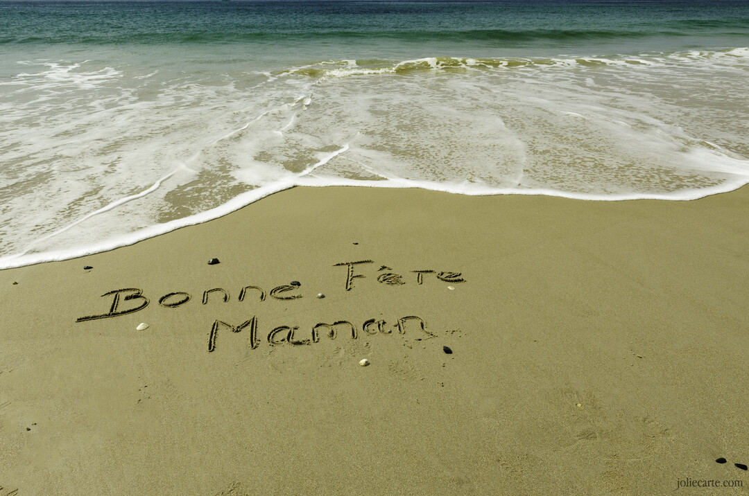 Le texte 'Bonne Fête Maman' est écrit à la main sur le sable humide d'une plage, avec la mer et son écume en arrière-plan.