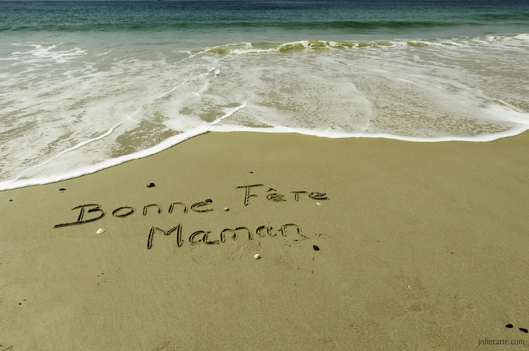 Le texte 'Bonne Fête Maman' est écrit à la main sur le sable humide d'une plage, avec la mer et son écume en arrière-plan.