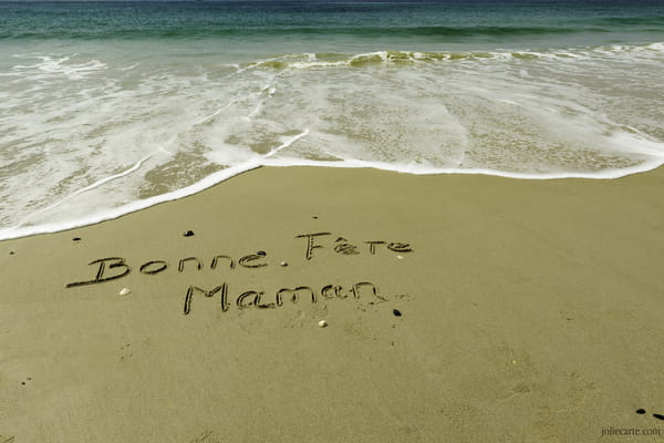 Carte pour la Fête des Mères avec le message 'Bonne Fête Maman' manuscrit sur le sable d'une plage, au bord des vagues de l'océan.