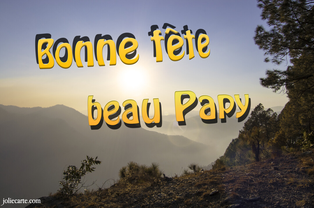 Un texte jaune en 3D 'Bonne fête beau Papy' flotte au-dessus d'un panorama de montagnes brumeuses, baignées dans la lumière chaude d'un soleil levant.