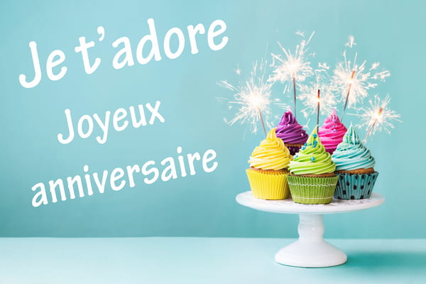 Carte d'anniversaire avec des cupcakes multicolores sur un présentoir, décorés de cierges magiques. Texte : 'Je t'adore, Joyeux anniversaire'.