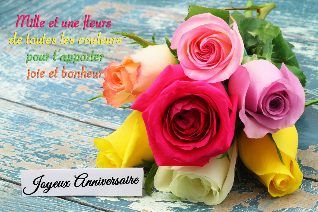 Un bouquet de roses multicolores (jaune, rose, fuchsia) repose sur une table en bois bleu rustique, avec une étiquette 'Joyeux Anniversaire'.
