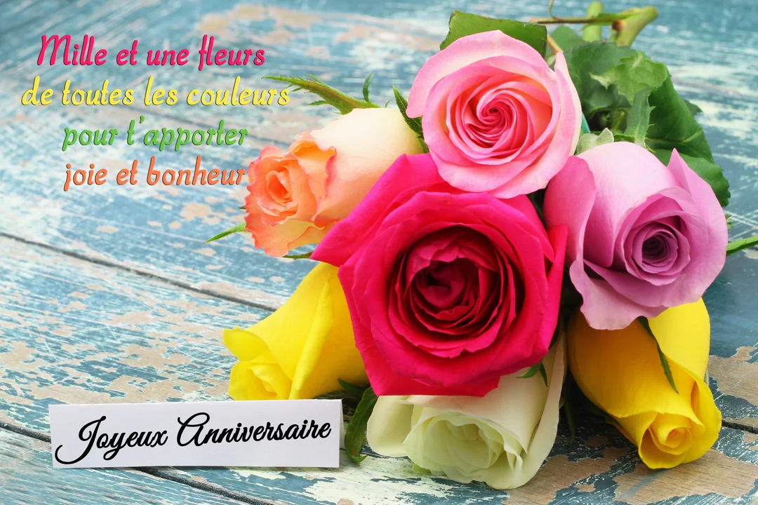 Un bouquet de roses multicolores (jaune, rose, fuchsia) repose sur une table en bois bleu rustique, avec une étiquette 'Joyeux Anniversaire'.