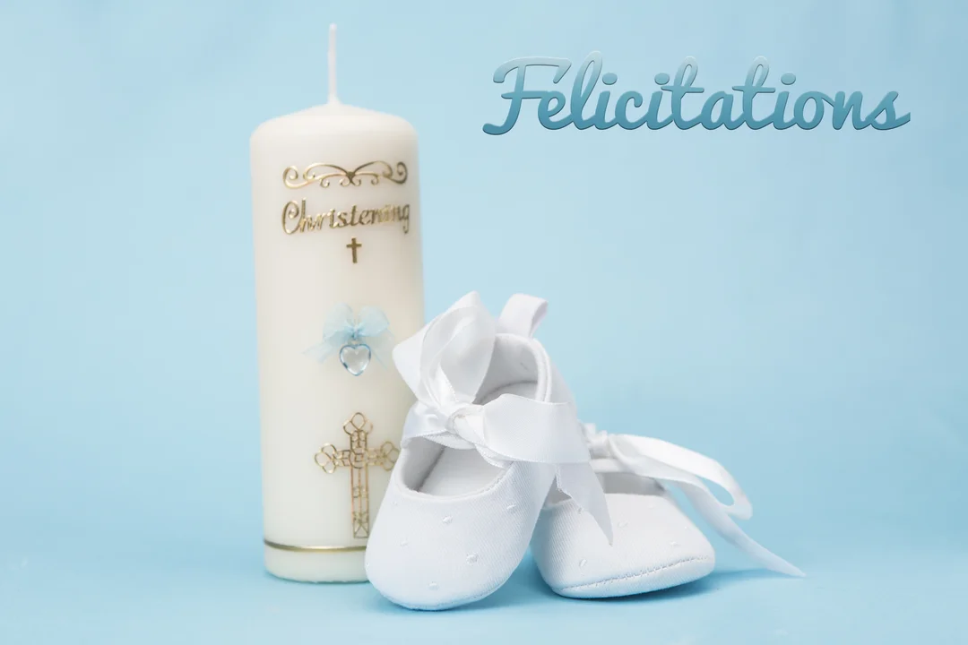 Une bougie de baptême blanche et dorée est posée à côté d'une paire de chaussons de bébé blancs sur un fond bleu clair.
