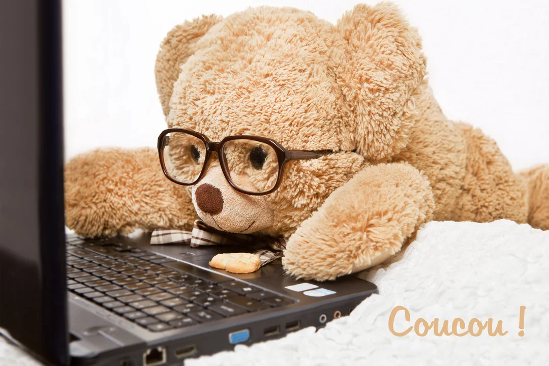 Un ours en peluche à lunettes est allongé confortablement dans un lit blanc, regardant l'écran d'un ordinateur portable posé devant lui.