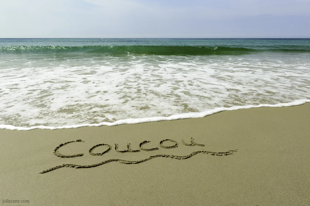 Sur une plage de sable fin, le mot 'Coucou' est dessiné près du rivage où une vague d'eau verte s'apprête à déferler.