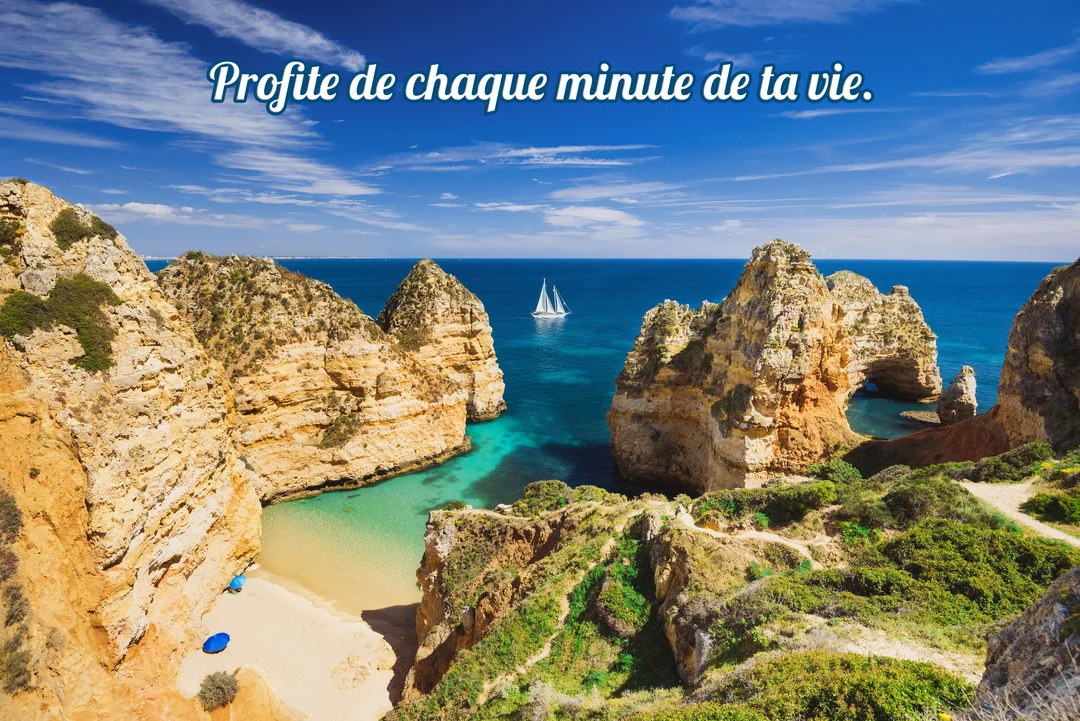 Vue ensoleillée sur une crique aux falaises ocres, une plage de sable fin, une mer turquoise et un voilier naviguant à l'horizon.