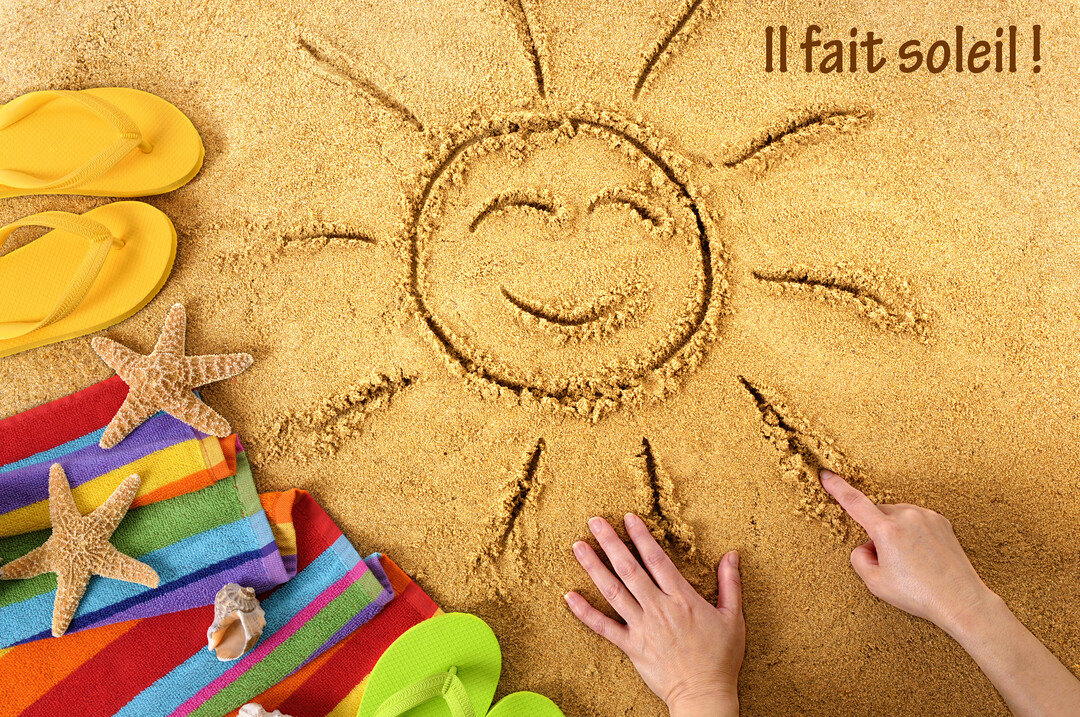 Vue de dessus d'une scène de plage où une main dessine un soleil souriant sur le sable, avec tongs et serviette multicolore.