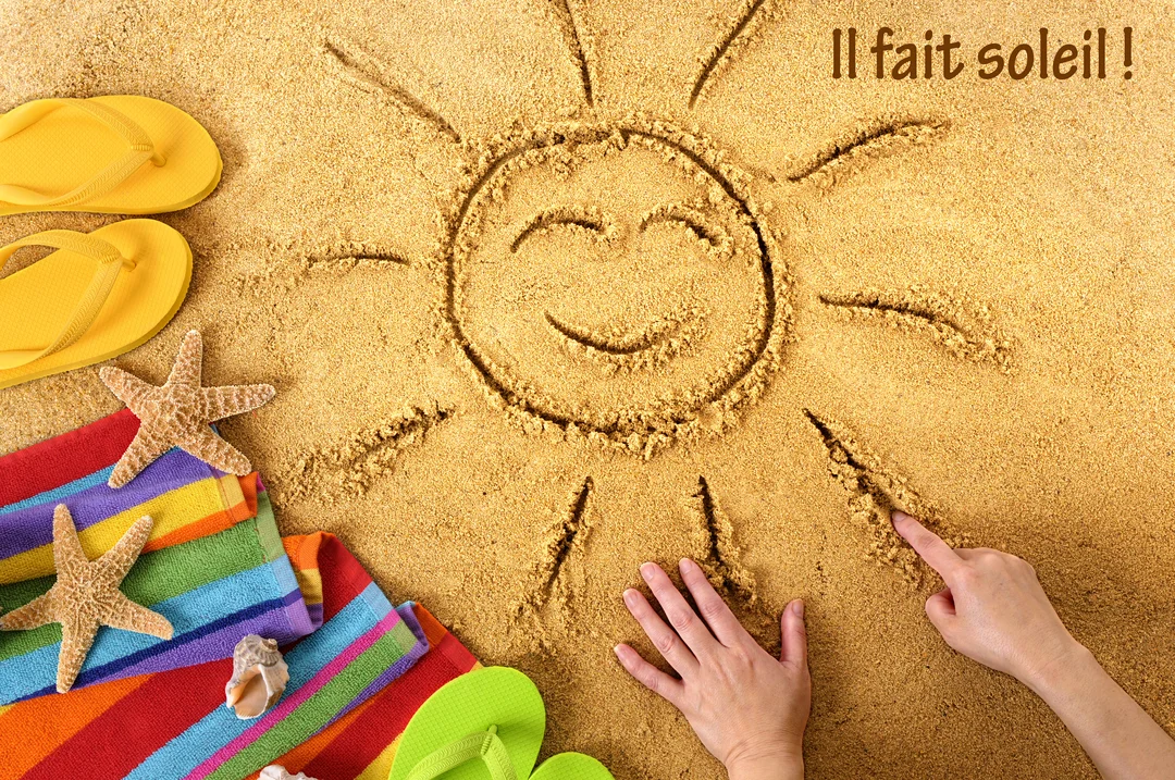 Vue de dessus d'une scène de plage où une main dessine un soleil souriant sur le sable, avec tongs et serviette multicolore.