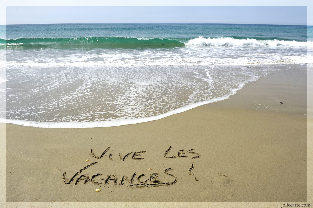 Photographie d'une plage où le message 'Vive les vacances !' est tracé sur le sable, face à une mer calme et un horizon clair.