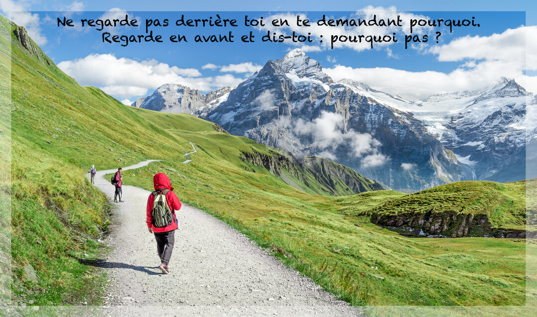 Une personne en anorak rouge marche sur un sentier de randonnée, s'éloignant à travers des collines verdoyantes vers de majestueuses montagnes enneigées.