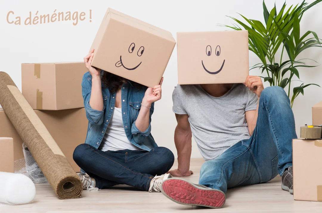 Un homme et une femme sont assis par terre au milieu de cartons, tenant chacun une boîte devant leur visage avec un smiley souriant.