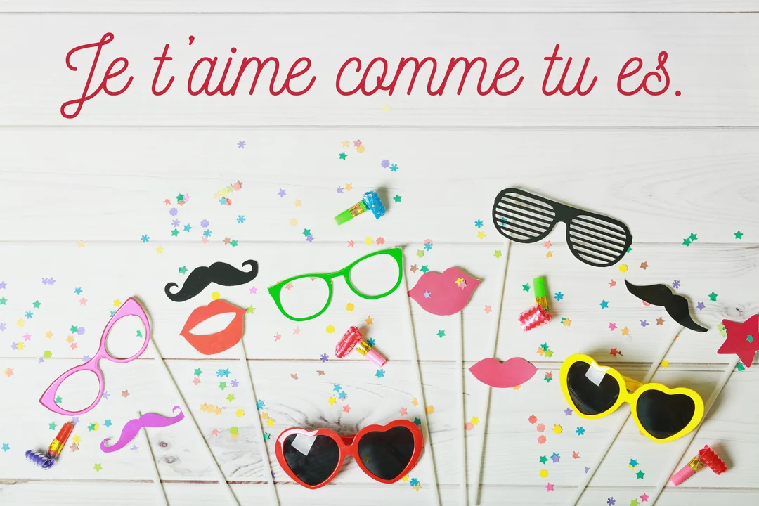 Une composition festive à plat d'accessoires de photobooth colorés sur un fond en bois blanc, avec le message d'amour manuscrit 'Je t'aime comme tu es' en rouge.