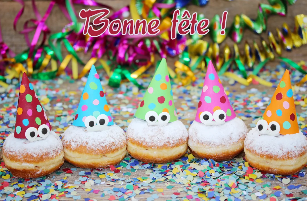 Une rangée de cinq beignets décorés de chapeaux pointus et d'yeux rigolos, posés sur une table en bois recouverte de confettis multicolores.