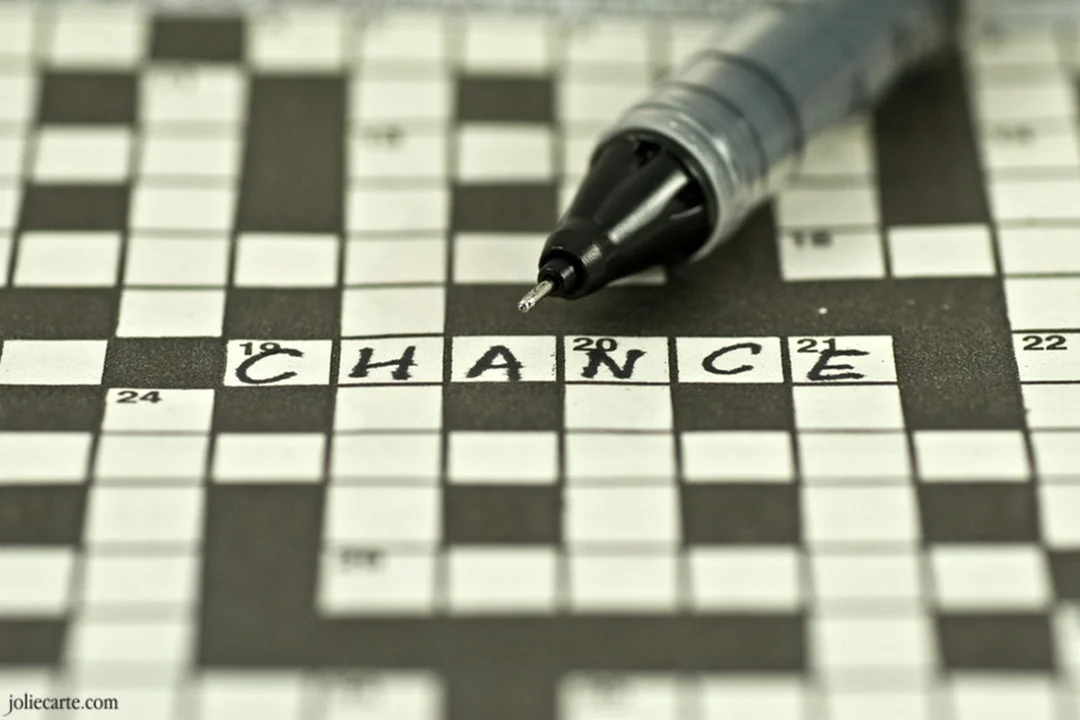Une photographie en gros plan montre le mot 'CHANCE' rempli sur une grille de mots croisés, avec la pointe d'un stylo juste au-dessus.