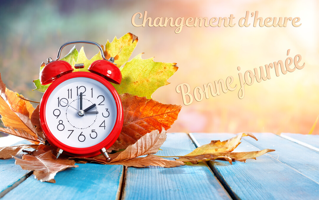 Un réveil rouge vintage est posé parmi des feuilles d'automne sur une surface en bois bleu, avec un fond lumineux et flou.