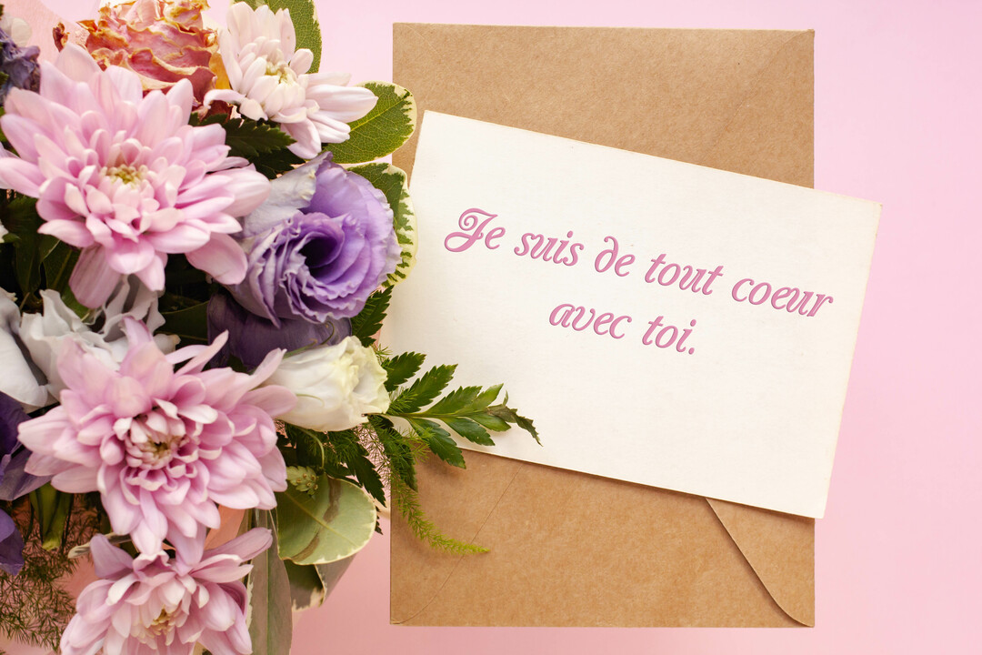 Un bouquet de fleurs roses et violettes est posé à côté d'une carte sur fond rose, avec un message de soutien manuscrit.