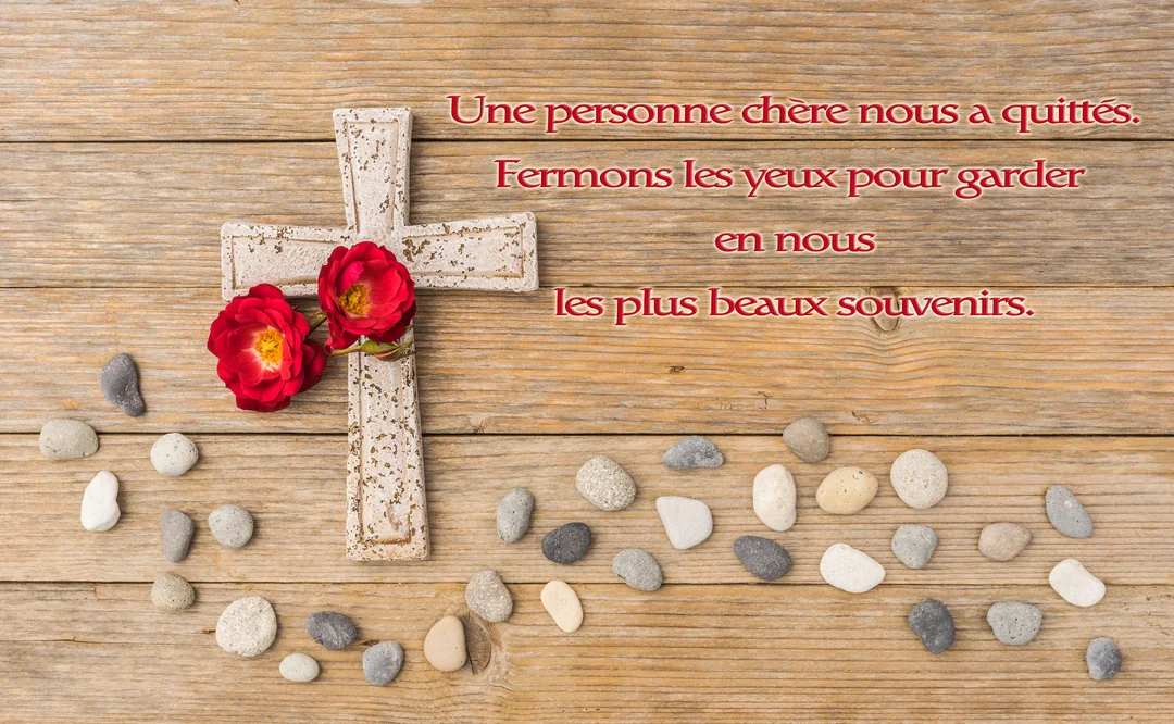 Sur un fond de planches de bois, une croix blanche ornée de deux roses rouges est posée à côté de galets.