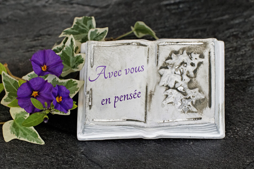 Une plaque en pierre blanche en forme de livre ouvert, gravée du texte "Avec vous en pensée", est posée sur une ardoise avec des fleurs violettes.
