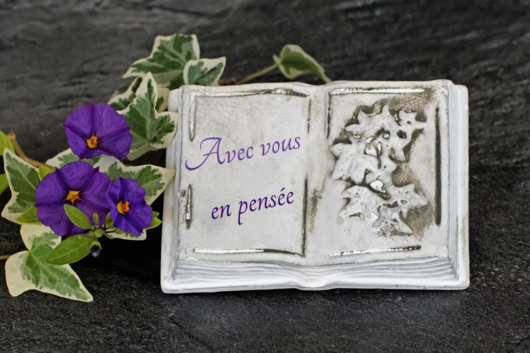 Une plaque en pierre blanche en forme de livre ouvert, gravée du texte "Avec vous en pensée", est posée sur une ardoise avec des fleurs violettes.