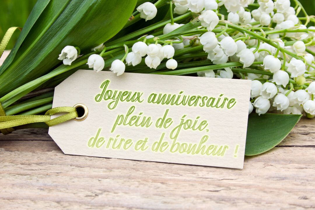 Un bouquet de muguet blanc et de feuilles vertes repose sur une surface en bois, avec une étiquette souhaitant un joyeux anniversaire.