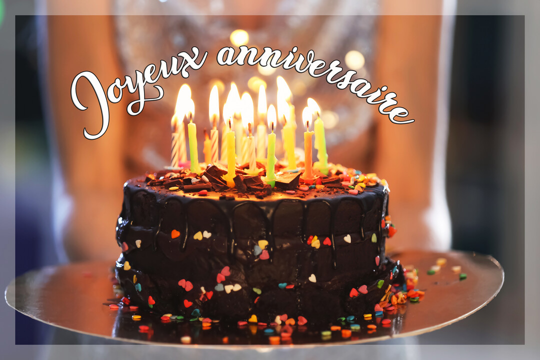 Une femme en arrière-plan flou présente un gâteau au chocolat sur un plateau, surmonté de nombreuses bougies colorées et allumées.