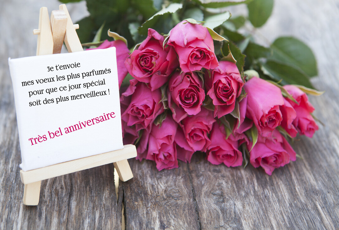 Un bouquet de roses fuchsia est posé sur une surface en bois à côté d'un petit chevalet affichant des vœux d'anniversaire manuscrits.