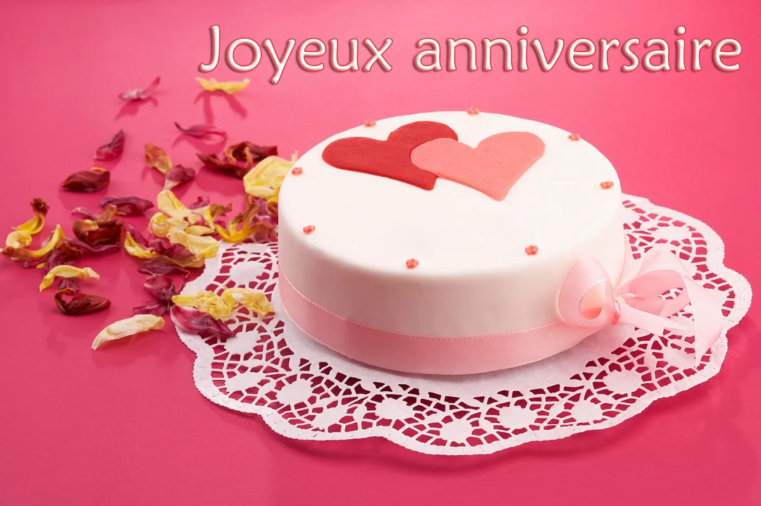 Un gâteau d'anniversaire blanc avec deux cœurs en sucre est posé sur un napperon, entouré de pétales, sur un fond rose.